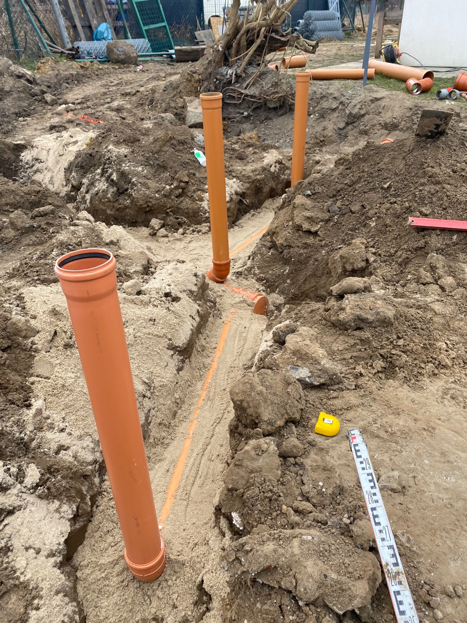 Wykop z pomarańczowymi rurami kanalizacyjnymi ułożonymi w piasku, widoczne narzędzia pomiarowe - poziomica i miarka.