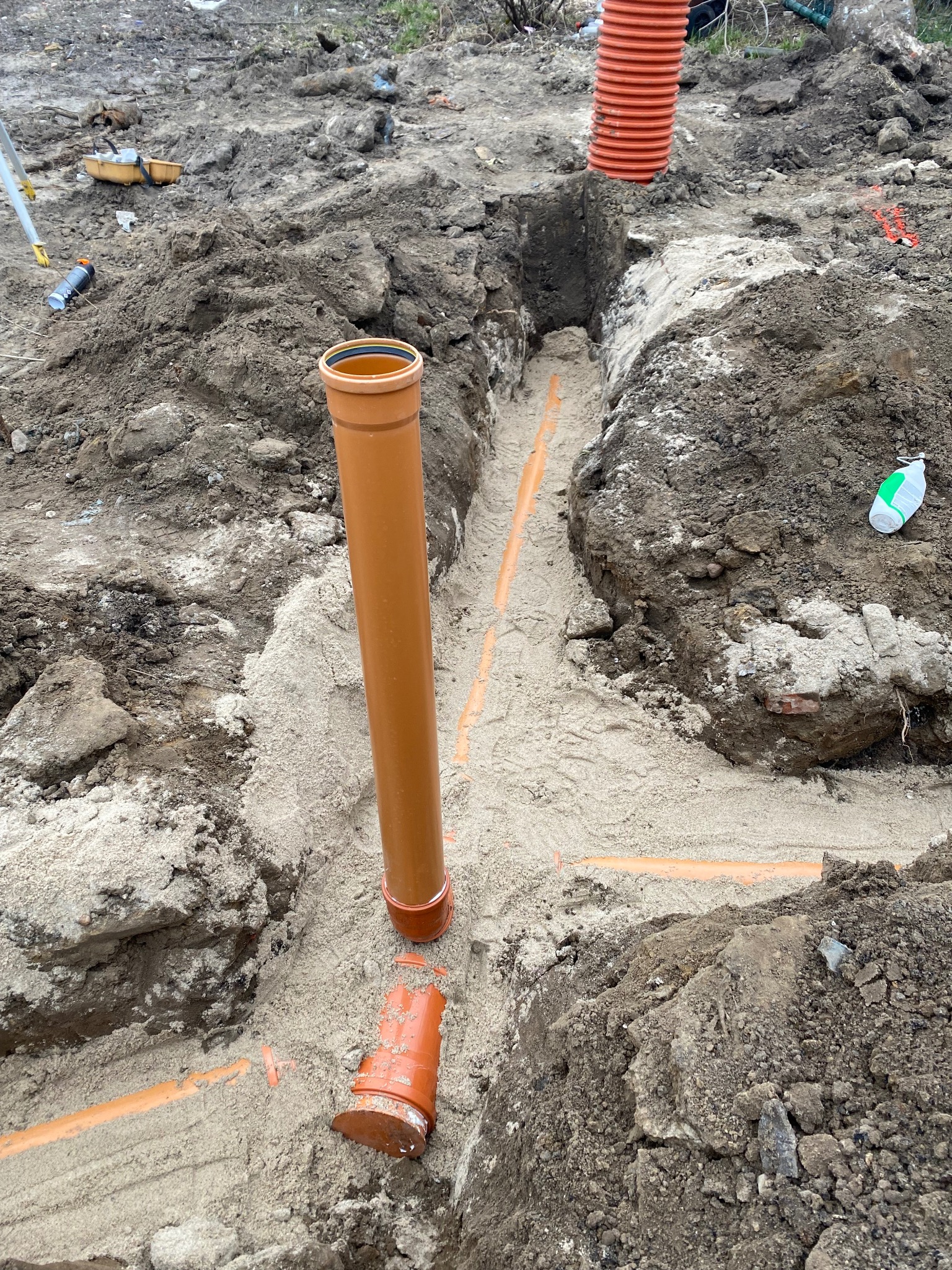 Wykop z pomarańczowymi rurami kanalizacyjnymi w piasku, widoczne elementy instalacji i narzędzia pomiarowe w tle.
