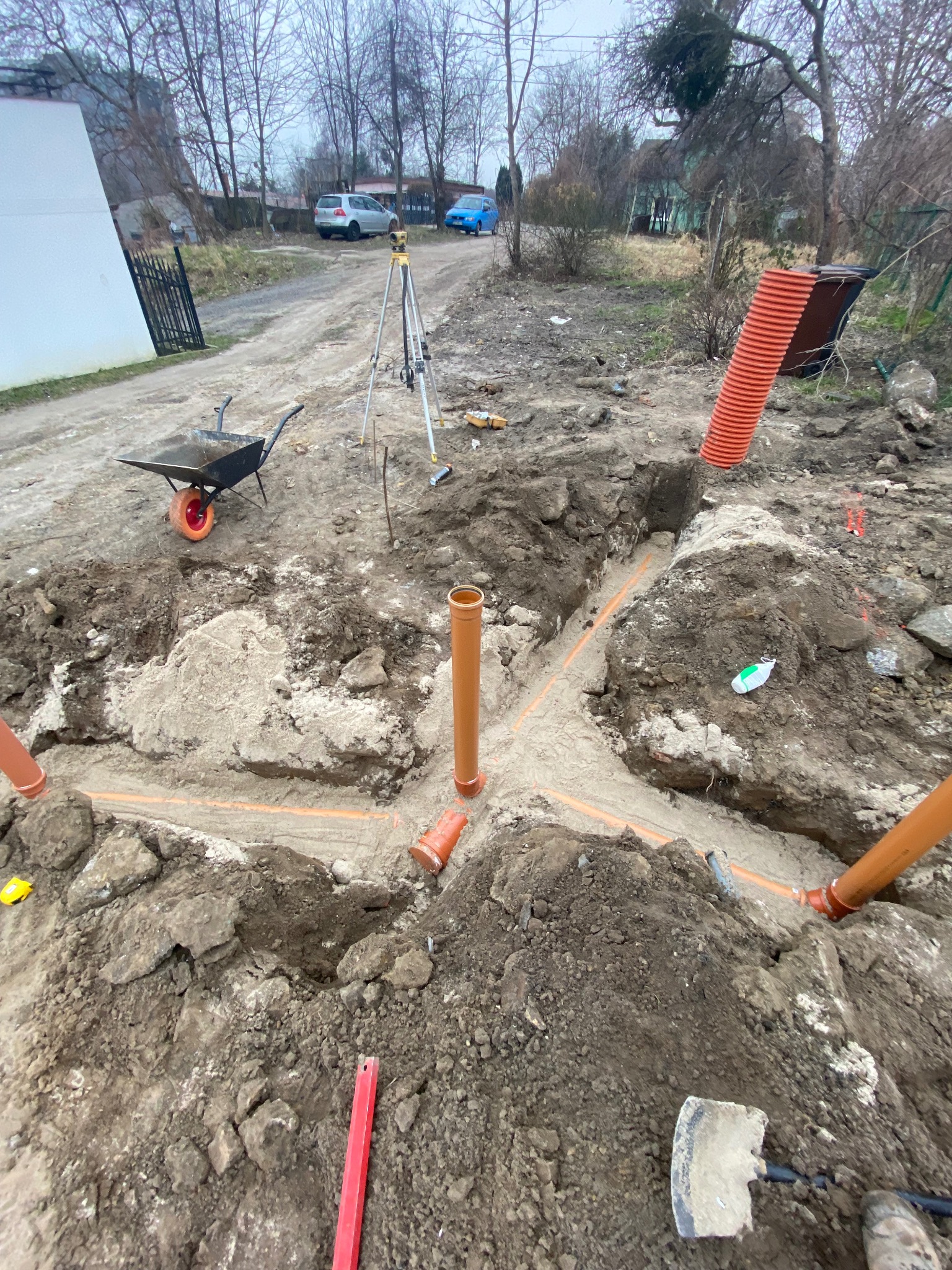 Wykonane wykopy pod instalację rur kanalizacyjnych z widocznymi pomarańczowymi rurami i narzędziami na placu budowy, w tle taczka i niwelator.