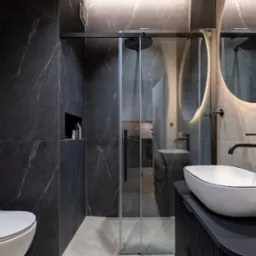 Nowoczesna łazienka z ciemnymi płytkami imitującymi marmur, kabina prysznicowa ze szklanymi drzwiami przesuwnymi, okrągłe lustro z podświetleniem LED i biała umywalka nablatowa z czarną armaturą.