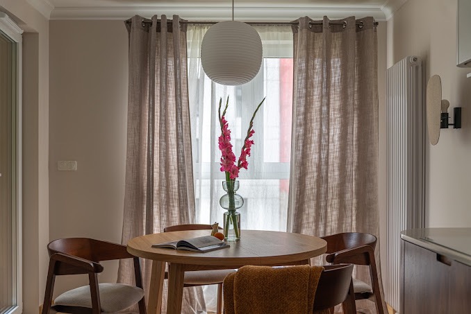 Jasne wnętrze jadalni z okrągłym drewnianym stołem, krzesłami z ciemnego drewna, lnianymi zasłonami, lampą kulą i wazonem z różowymi mieczykami na tle okna.