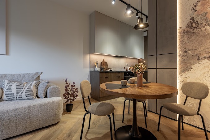 Nowoczesne wnętrze: jasna kuchnia z drewnianym blatem i szarymi szafkami, okrągły stół z krzesłami na tle ściany z efektem kamienia, oświetlenie punktowe i drewniana podłoga ułożona w jodełkę.