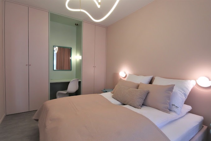 Pokój w pastelowych odcieniach z łóżkiem, szafą w zabudowie z biurkiem i lustrem, oraz lampą sufitową w kształcie neonowej linii.