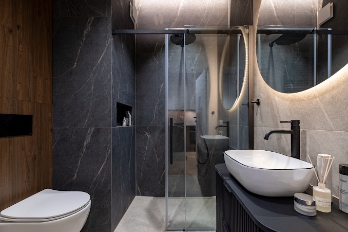 Nowoczesna łazienka z ciemnymi płytkami imitującymi marmur, kabina prysznicowa ze szklanymi drzwiami przesuwnymi, okrągłe lustro z podświetleniem LED i biała umywalka nablatowa z czarną armaturą.