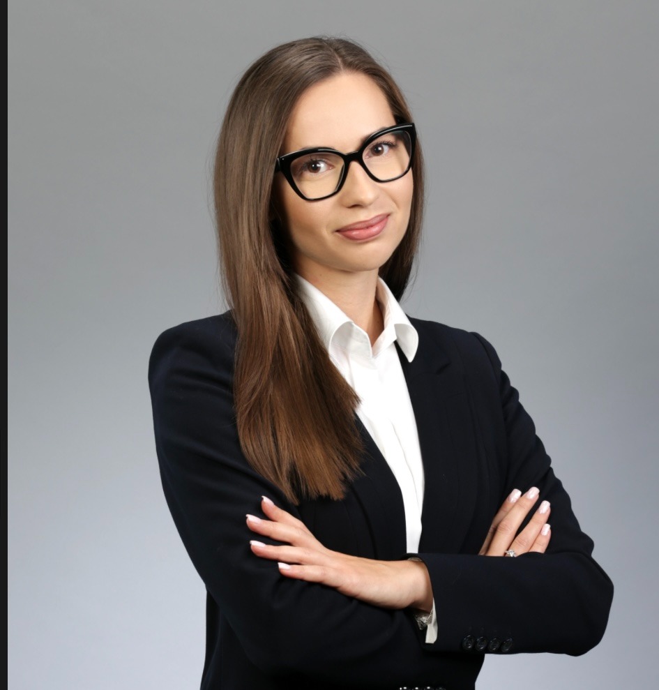 Portret kobiety w czarnym garniturze i okularach, z założonymi rękami na szarym tle.