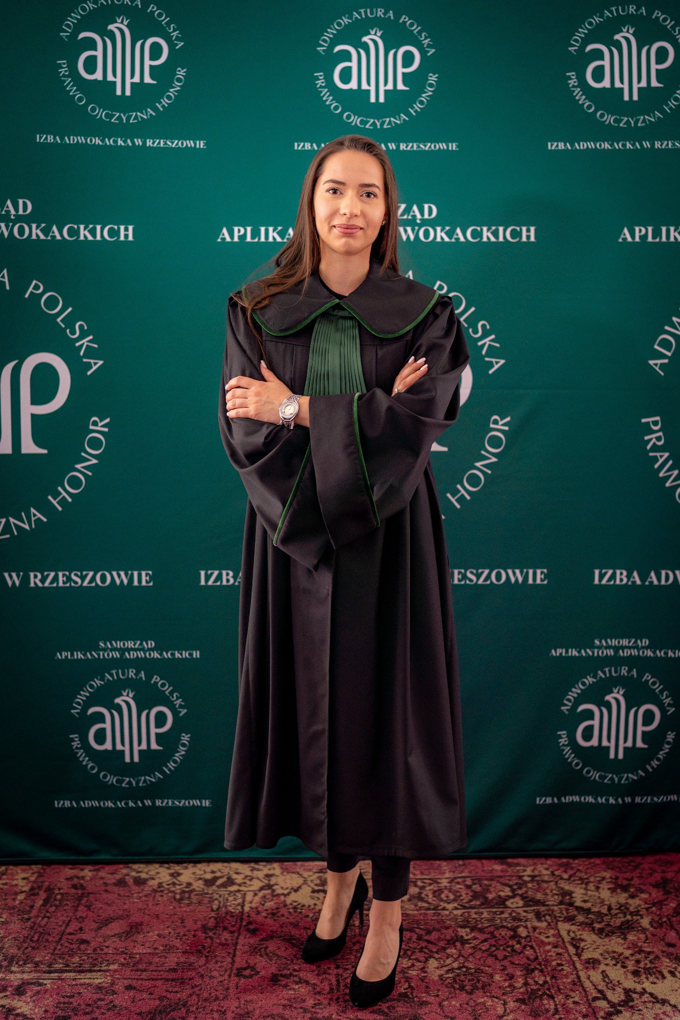 Portret młodej kobiety w todze adwokackiej, na tle z logo Izby Adwokackiej w Rzeszowie, z zegarkiem na nadgarstku i czarnych szpilkach.