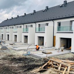 WIKING PRO sp. z o.o. - Budowa szeregu nowoczesnych domów jednorodzinnych z płaskimi dachami, widoczne prace brukarskie przed wejściami, pracownik w pomarańczowej kamizelce, gruz i materiały budowlane na pierwszym planie.