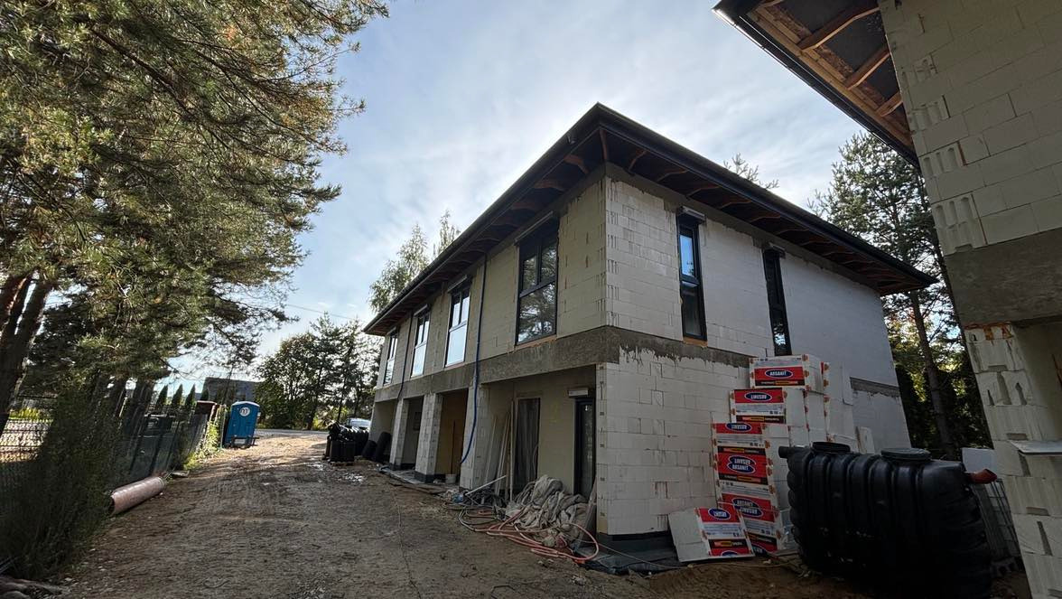 Wznoszenie dwukondygnacyjnego budynku szeregowego z białych bloczków, widoczne okna wstawione na parterze i piętrze, na placu budowy składowane materiały budowlane i zbiornik na deszczówkę, w tle...