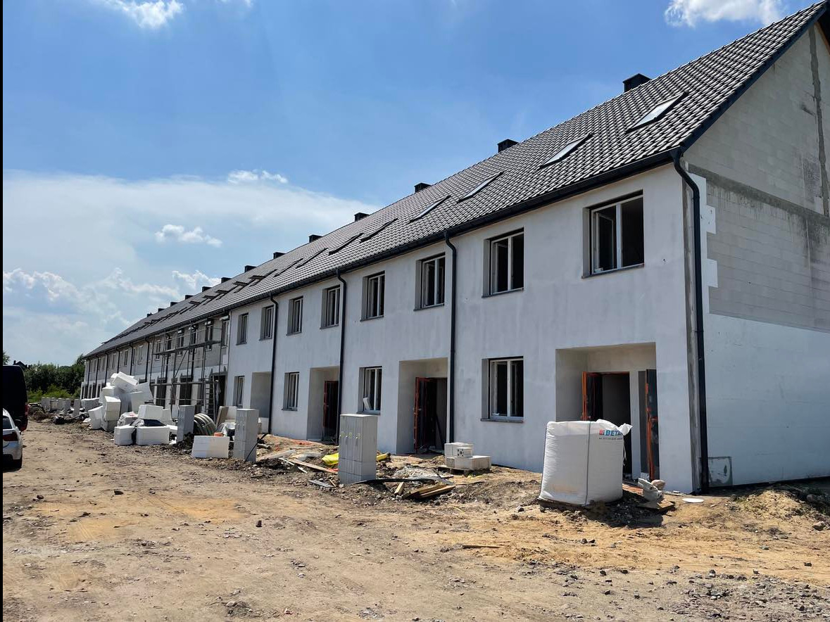 Rząd nowych, szeregowych domów w trakcie budowy, z widocznymi materiałami budowlanymi na placu, w słoneczny dzień.