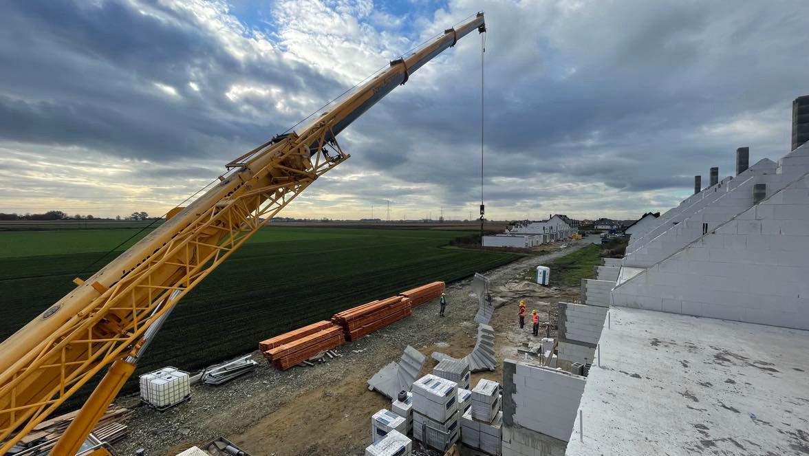 Żuraw wieżowy na placu budowy domów szeregowych z białych bloczków, widoczne składowane drewno i pracownicy w kaskach na tle pochmurnego nieba.