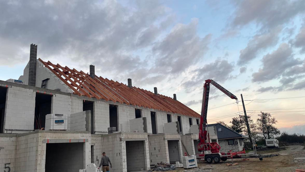 Budowa szeregu domów jednorodzinnych w stanie surowym otwartym, widoczne bloczki z betonu komórkowego, drewniana konstrukcja dachu, dźwig budowlany w tle, pochmurne niebo.