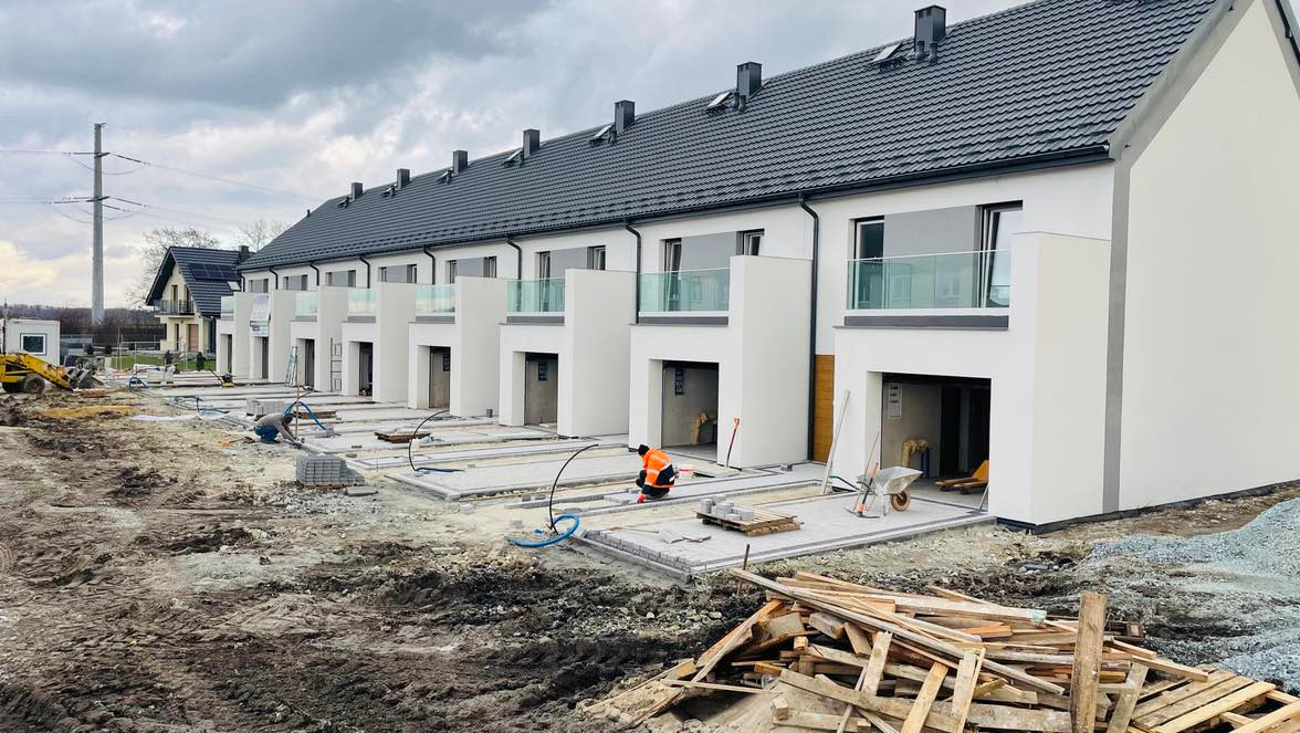 Budowa szeregu nowoczesnych domów jednorodzinnych z płaskimi dachami, widoczne prace brukarskie przed wejściami, pracownik w pomarańczowej kamizelce, gruz i materiały budowlane na pierwszym planie.