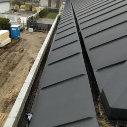 Gryfdach - Nowy, grafitowy dach z blachy na budynku w trakcie budowy. Widoczne rynny i elementy konstrukcyjne. W tle zabudowa jednorodzinna i drzewa bez liści.