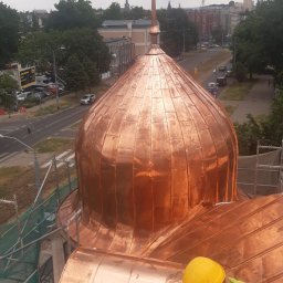 Gryfdach - Miedziana kopuła budynku sakralnego w trakcie renowacji, widok z góry na pracownika w żółtym kasku, miasto w tle.