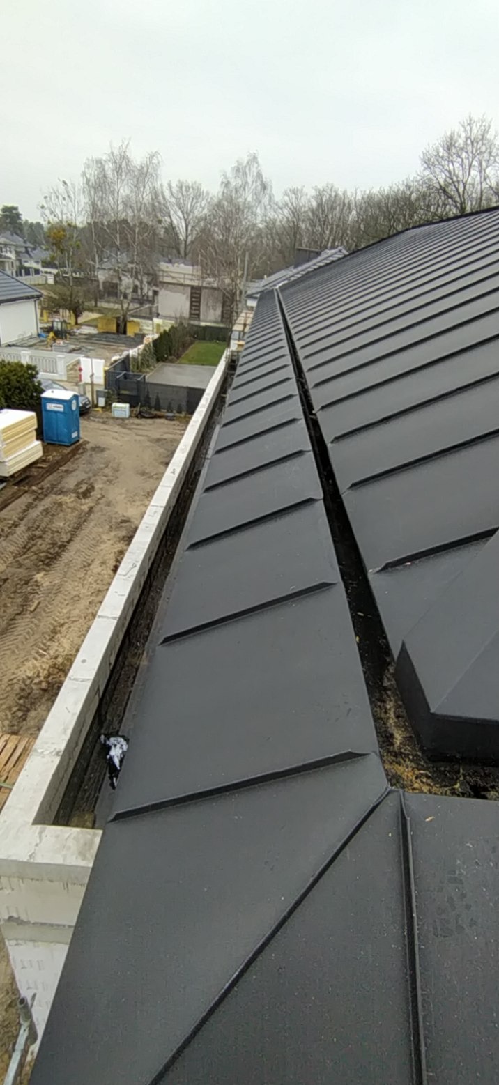 Nowy, grafitowy dach z blachy na budynku w trakcie budowy. Widoczne rynny i elementy konstrukcyjne. W tle zabudowa jednorodzinna i drzewa bez liści.