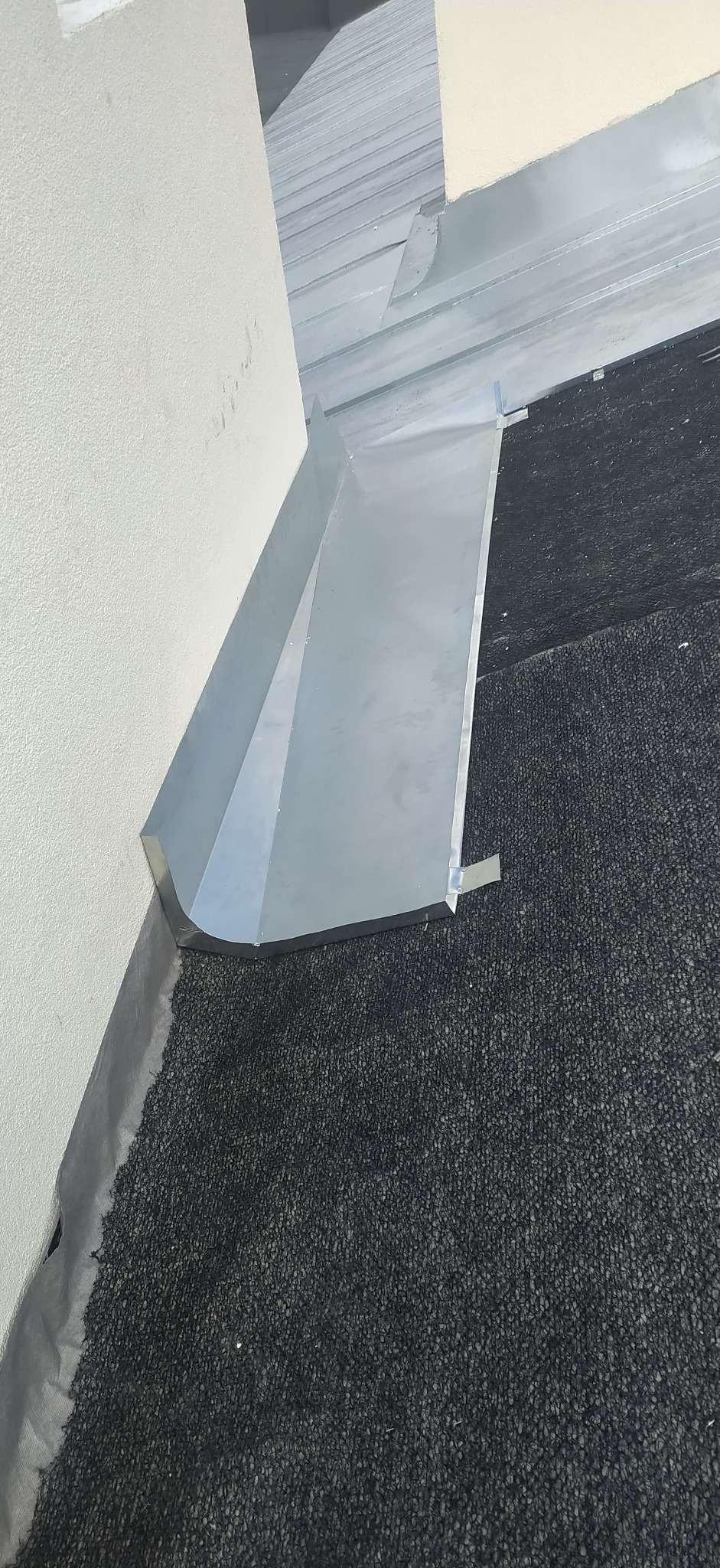 Szczegółowe ujęcie nowej, błyszczącej, metalowej obróbki blacharskiej dachu, przylegającej do ściany. Widoczna czarna papa i fragment blaszanej części dachu.