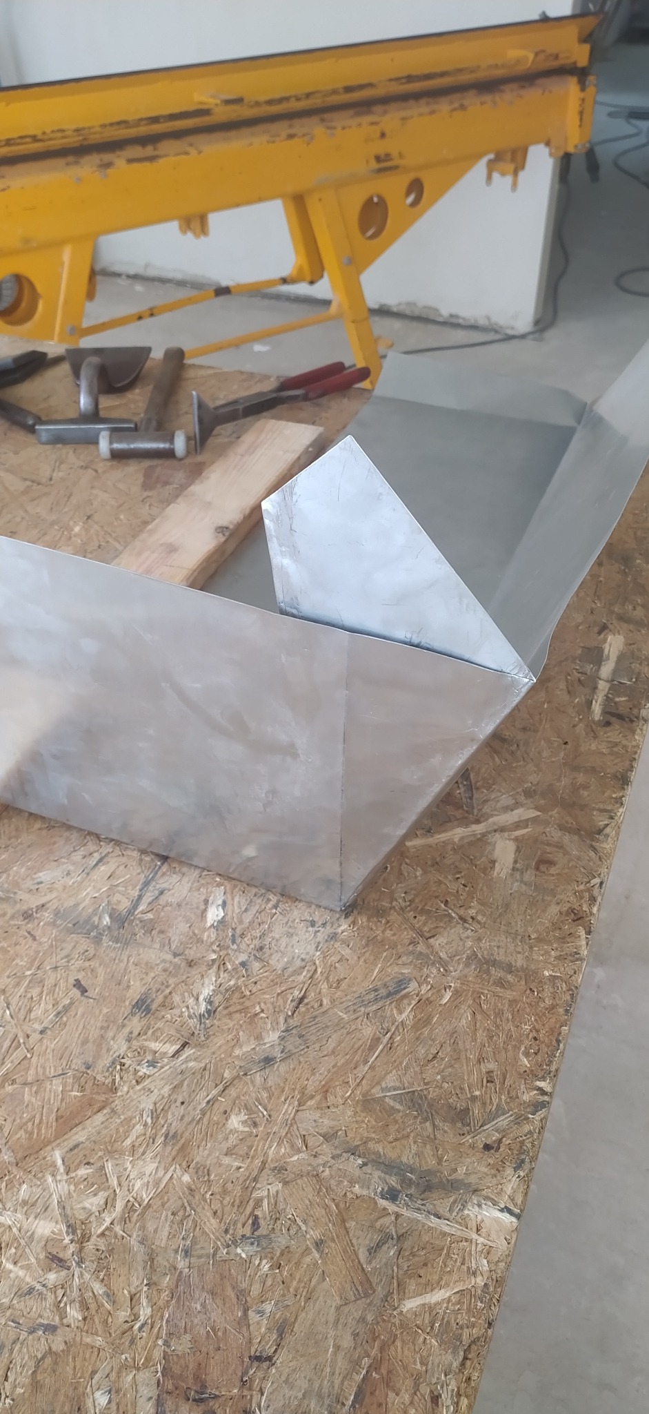 Metalowa obróbka blacharska, zagięta pod kątem, leży na stole warsztatowym obok narzędzi dekarskich i żółtej zaginarki do blachy. Ukazany proces formowania blachy.