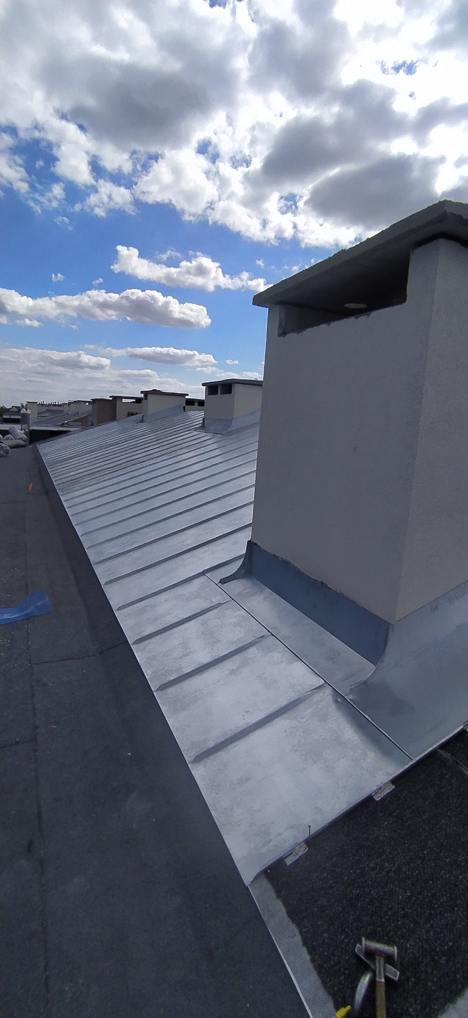 Metalowy dach z kominem w słoneczny dzień. Widoczne narzędzia blacharskie. Nowe pokrycie dachu z blachy na tle błękitnego nieba z chmurami.