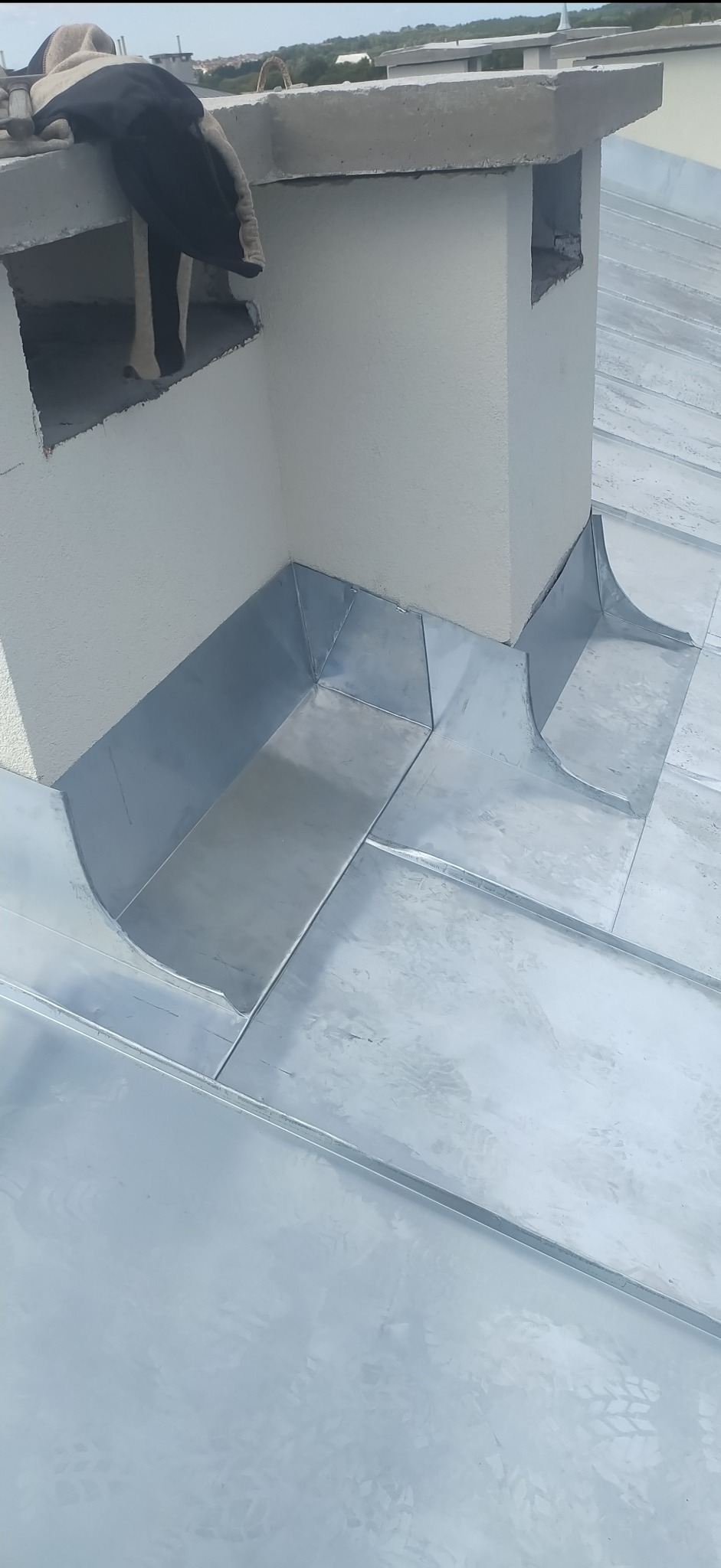 Szczegółowe ujęcie blaszanych obróbek komina na dachu, z widocznymi łączeniami i elementami mocującymi, wykonane w jasnym oświetleniu.