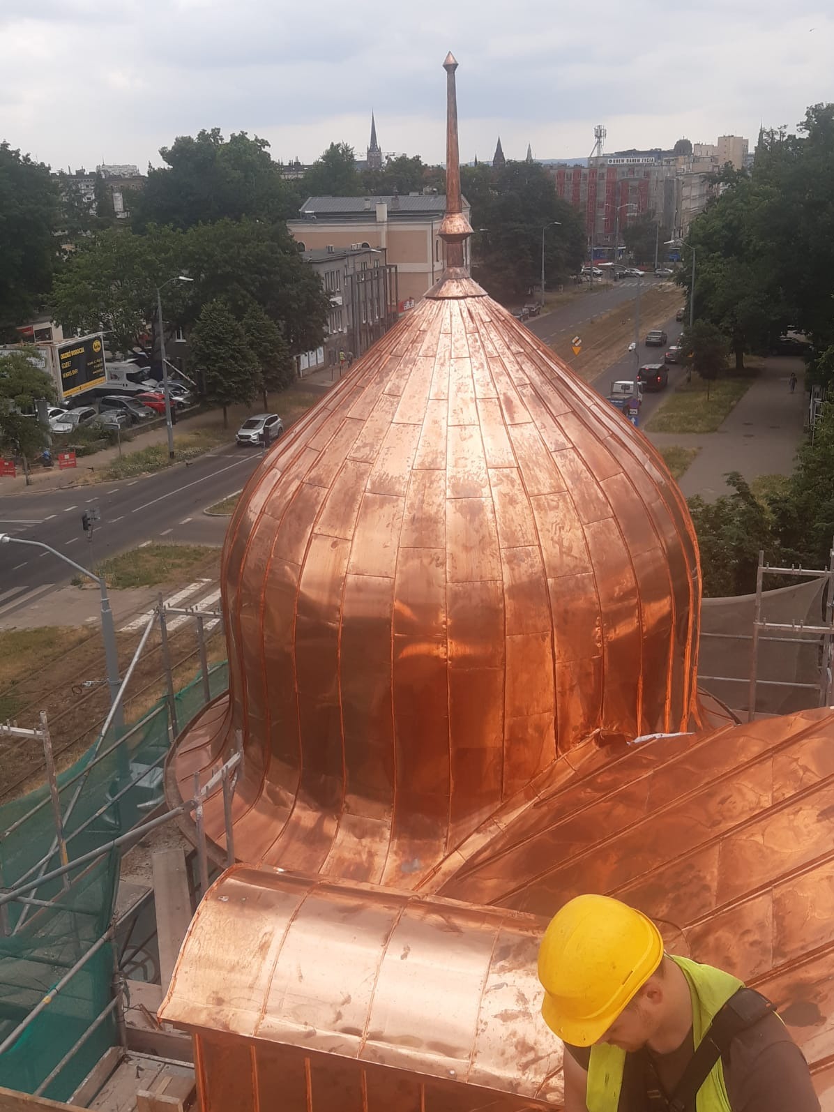 Miedziana kopuła budynku sakralnego w trakcie renowacji, widok z góry na pracownika w żółtym kasku, miasto w tle.