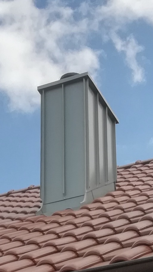 Szary, metalowy komin na dachu z czerwonej dachówki, na tle błękitnego nieba z chmurami.