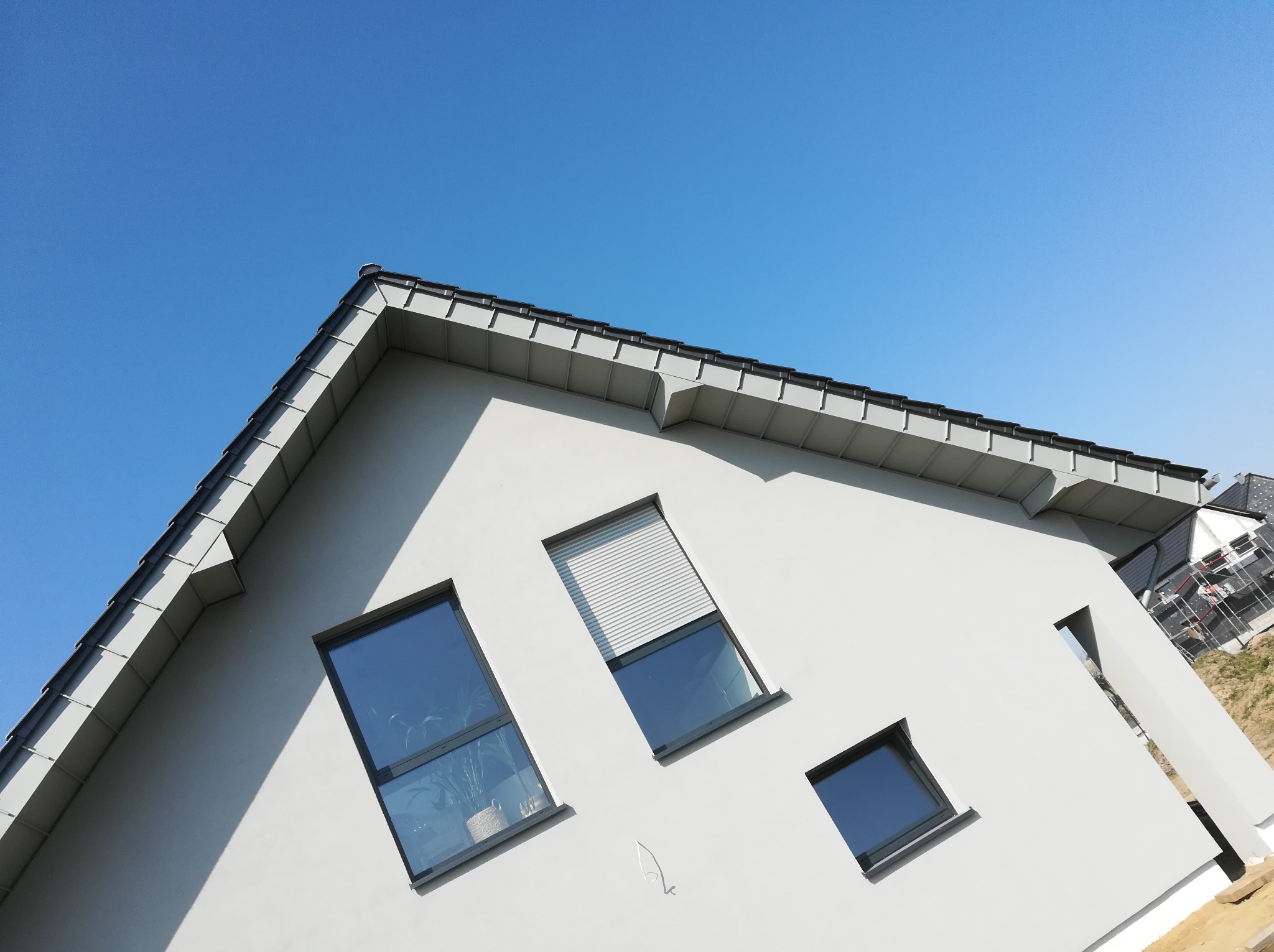 Nowoczesny, jasnoszary dom jednorodzinny z ciemną dachówką i oknami o różnych rozmiarach, sfotografowany pod kątem z dołu na tle błękitnego nieba.