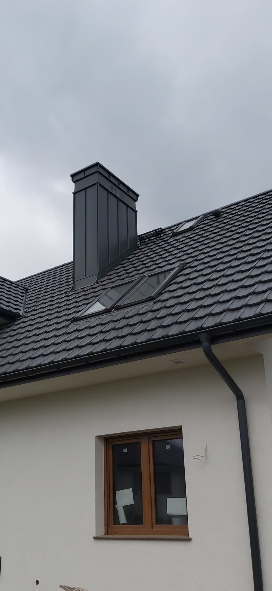 Nowoczesny dach z dachówką ceramiczną w kolorze grafitowym, z oknami połaciowymi i obróbką komina z blachy. Widoczny fragment elewacji budynku z oknem.