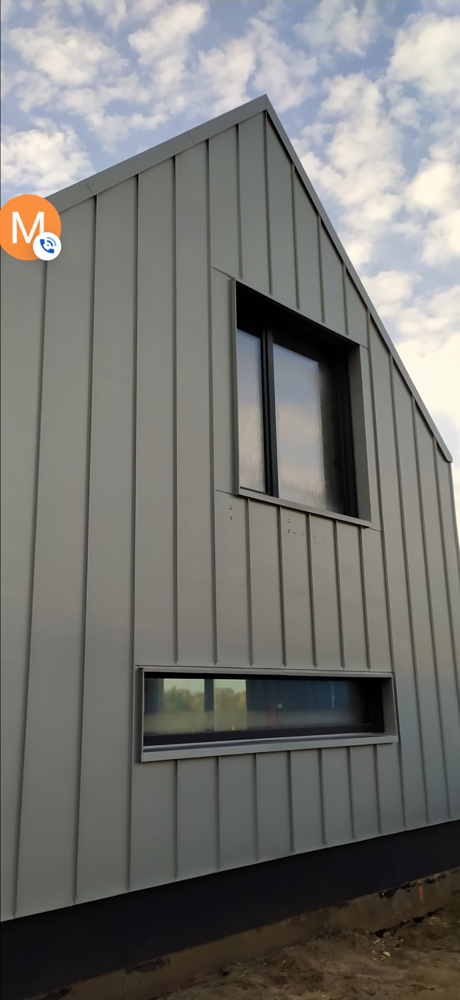 Nowoczesny dom jednorodzinny z szarą elewacją z blachy trapezowej i minimalistycznymi oknami, widok z dołu na tle błękitnego nieba.