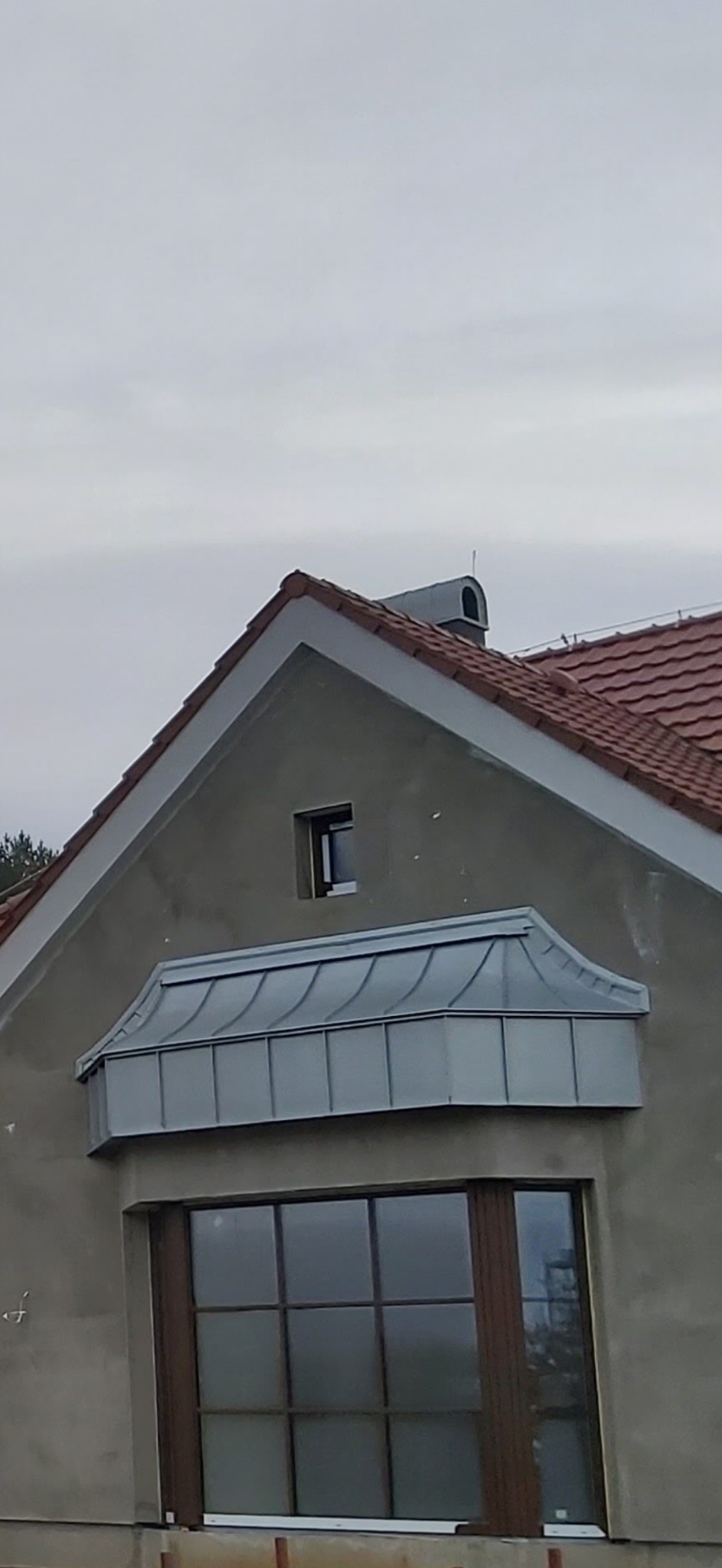 Szara fasada domu z oknami, wykończona blachą w kształcie falistym nad oknem. Czerwony dach z kominem w tle, pochmurne niebo.
