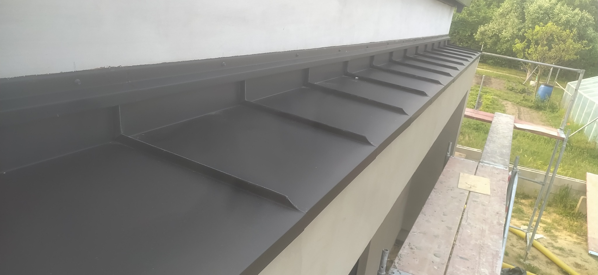 Nowoczesne, czarne obróbki blacharskie na dachu budynku, widoczne z rusztowania. Kąt ujęcia podkreśla geometrię i precyzję wykonania.