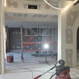KD Reno Build - Wnętrze w trakcie szpachlowania i wygładzania ścian, widoczne rusztowanie, szpachlowane ściany, lampa budowlana i odkurzacz przemysłowy.