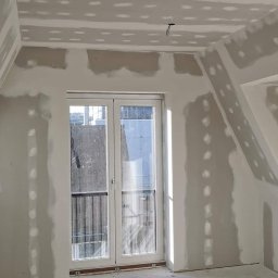 KD Reno Build - Pokój w trakcie przygotowania ścian i sufitu pod malowanie, widoczne miejsca po szpachlowaniu i wygładzaniu, okno balkonowe z widokiem na budynek z balkonami.