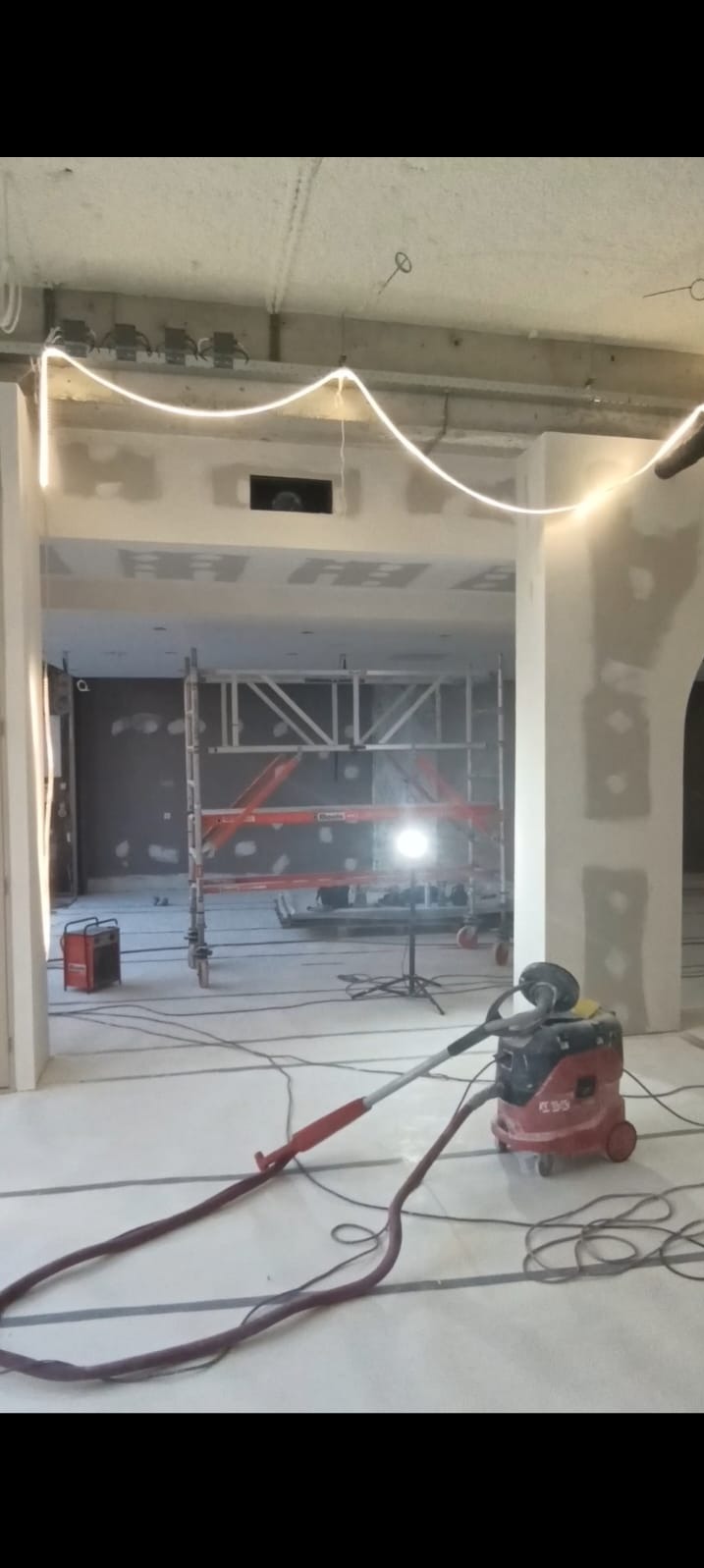 Wnętrze w trakcie szpachlowania i wygładzania ścian, widoczne rusztowanie, szpachlowane ściany, lampa budowlana i odkurzacz przemysłowy.