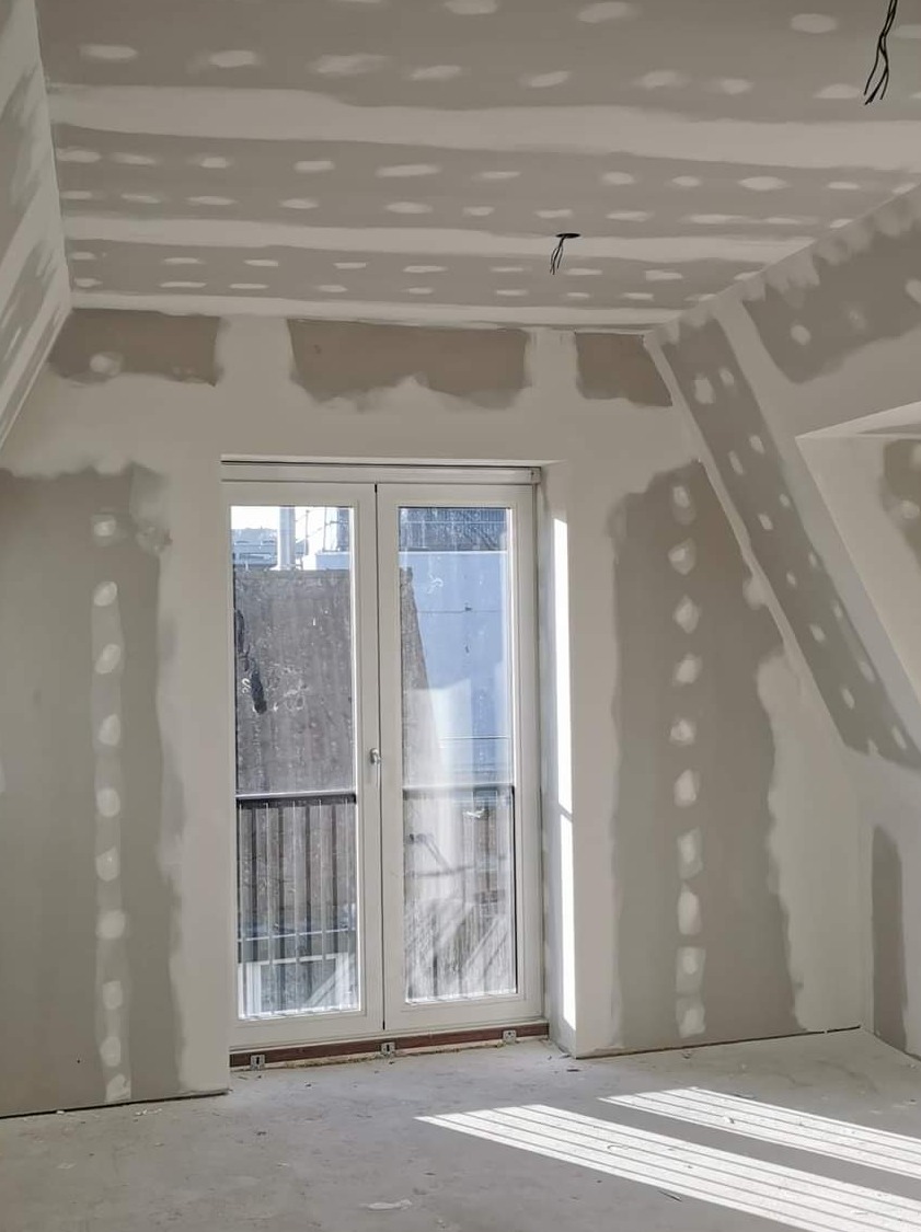 Pokój w trakcie przygotowania ścian i sufitu pod malowanie, widoczne miejsca po szpachlowaniu i wygładzaniu, okno balkonowe z widokiem na budynek z balkonami.