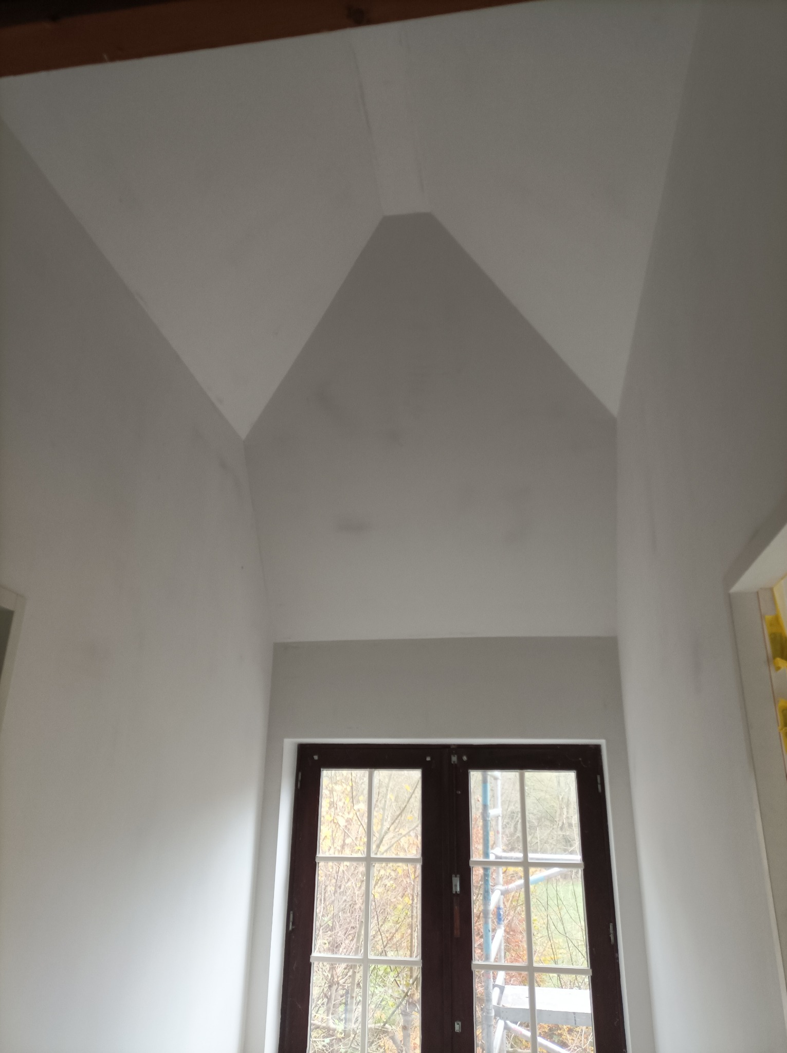 Surowe ściany i sufit poddasza po wstępnym wykończeniu w Garkach, widoczne ślady po szpachlowaniu i przygotowaniu pod malowanie, okno z widokiem na zewnątrz i fragment rusztowania.