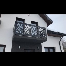 Dekory z metalu - Czarny, metalowy balkon z dekoracyjnym, laserowo wycinanym wzorem liści na tle białej ściany domu z ciemnymi oknami i szarym, kamiennym wykończeniem.