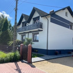 Dekory z metalu - Nowoczesny dom z białymi ścianami i dekoracyjnym, metalowym balkonem z motywem roślinnym, osoba opiera się o barierkę balkonu, widok z zewnątrz na podwórze z kostką brukową i ogrodzeniem.