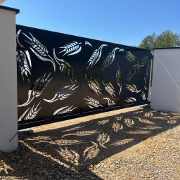 Dekory z metalu - Nowoczesna, przesuwna brama wjazdowa z motywem roślinnym, rzucająca cienie na żwirowe podłoże w słoneczny dzień. Czarna brama z wycinanymi wzorami.