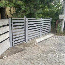Dekory z metalu - Otwarta, szara brama wjazdowa z poziomymi szczeblami, betonowy słupek imitujący drewno, kostka brukowa, zieleń w tle. Widok z zewnątrz posesji.