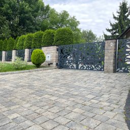 Dekory z metalu - Nowoczesna, grafitowa brama wjazdowa z laserowo ciętym wzorem roślinnym, osadzona na murowanych słupkach. W tle żywopłot z tuj i kostka brukowa na podjeździe.