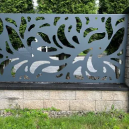 Nowoczesne ogrodzenie metalowe z dekoracyjnym wzorem wyciętym laserowo, osadzone na kamiennej podmurówce, z zielenią w tle.