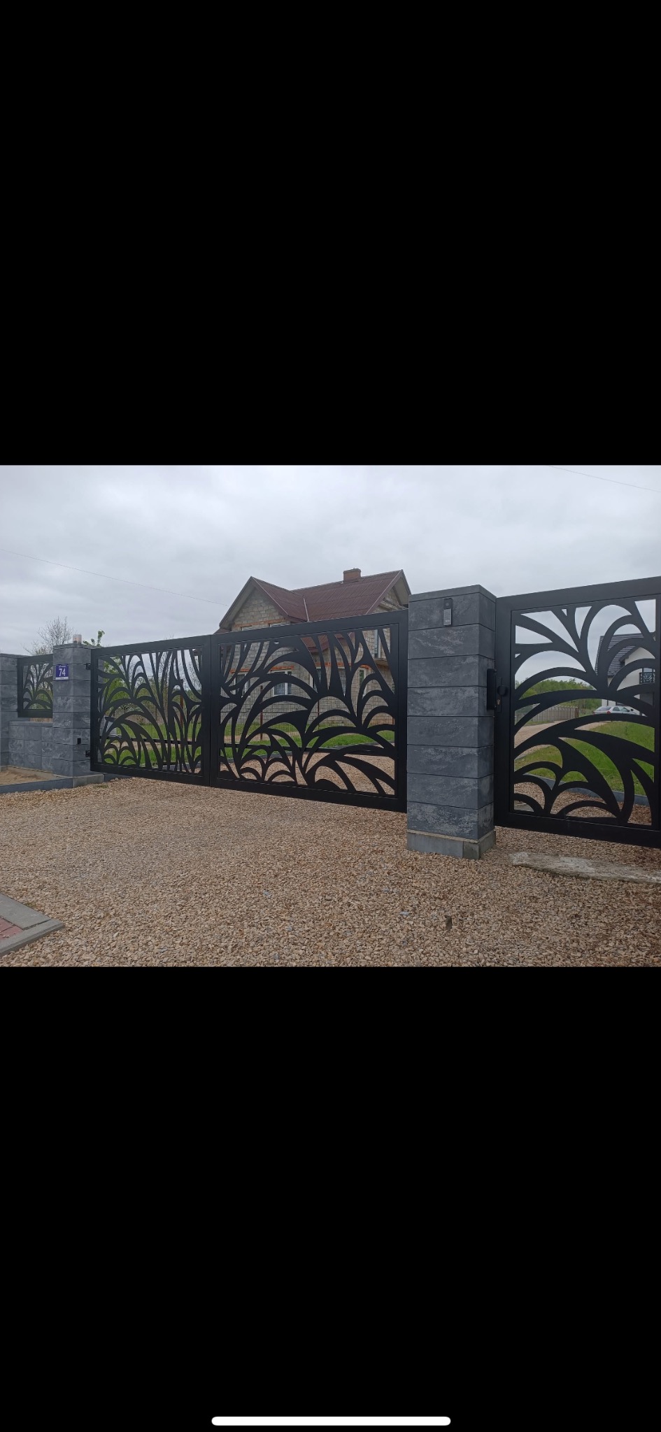 Nowoczesna, czarna brama wjazdowa z ażurowym wzorem liści, osadzona na szarych, kamiennych słupkach, prowadząca na żwirowy podjazd przed domem.