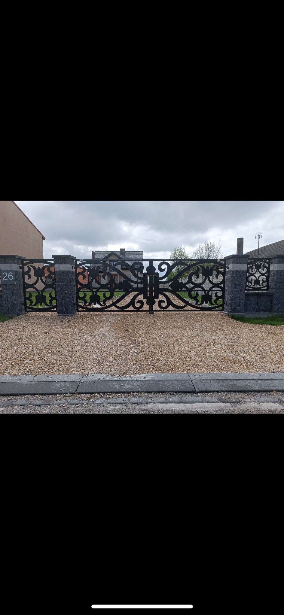 Dwuskrzydłowa, czarna brama z ornamentami, zamontowana na murowanych, szarych słupkach z numerem domu i dekoracyjnym panelem, prowadząca na żwirowy podjazd.