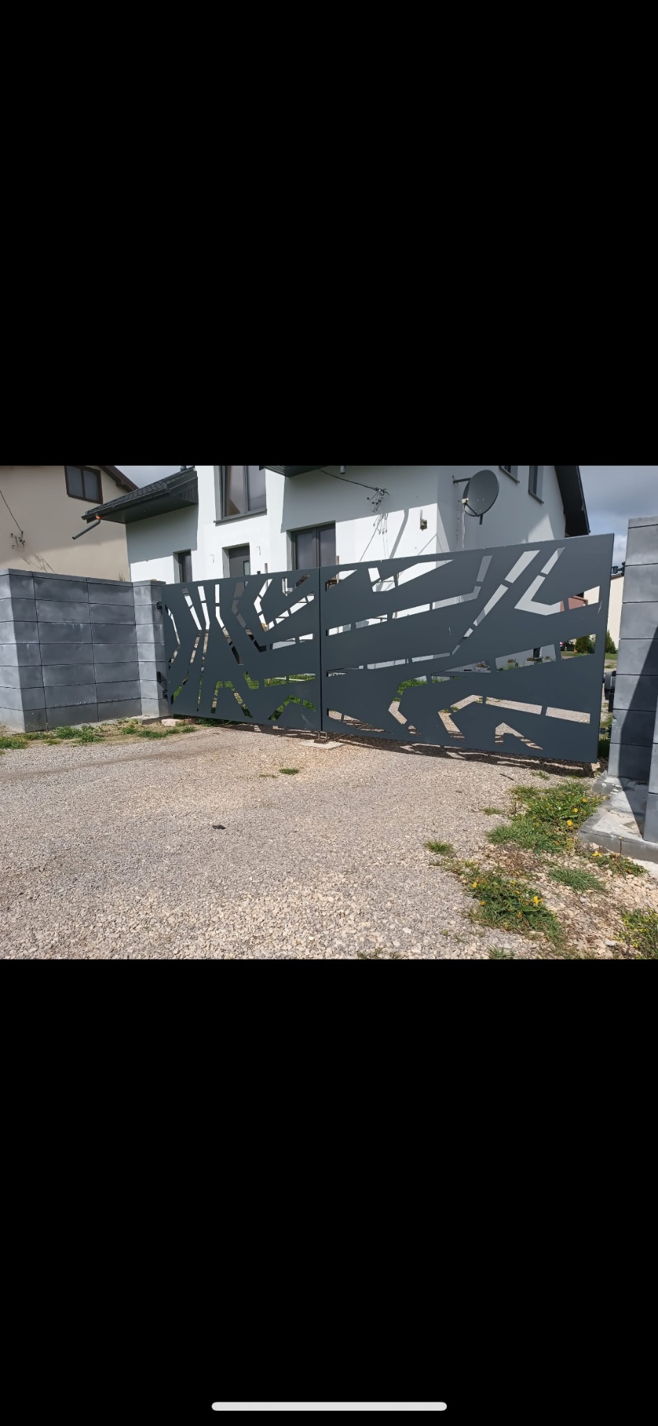 Nowoczesna, metalowa brama wjazdowa z abstrakcyjnym wzorem wycinanym laserowo, zamontowana na posesji z szarą kostką brukową i białym domem w tle.