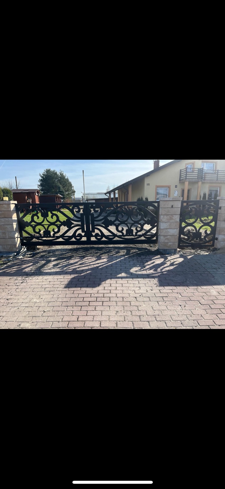 Czarna, ażurowa brama wjazdowa z ozdobnymi wzorami, osadzona na słupkach z jasnego kamienia, na tle domu jednorodzinnego i kostki brukowej.