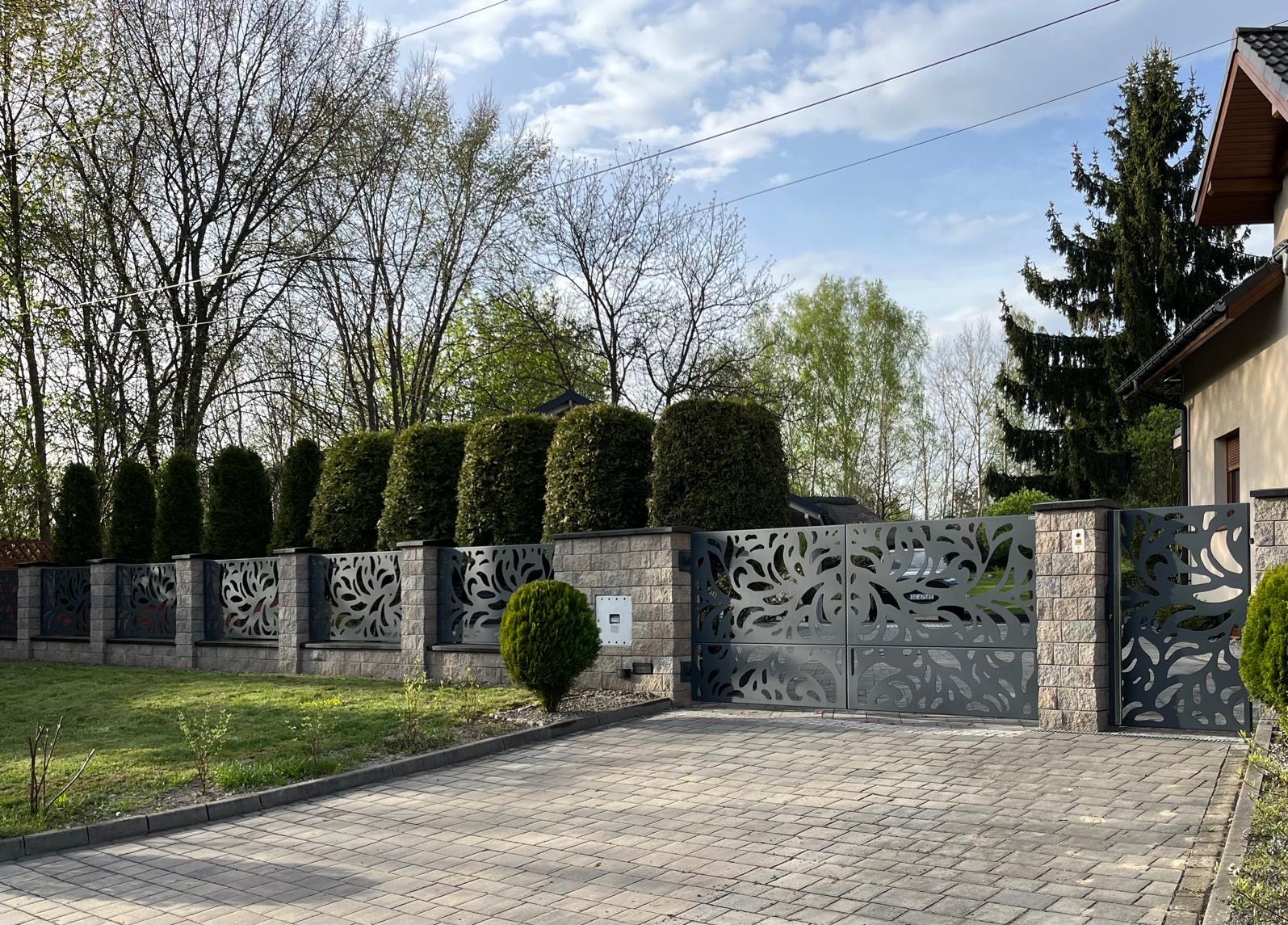 Nowoczesna, ażurowa brama dwuskrzydłowa i ogrodzenie panelowe z motywem roślinnym, osadzone na murowanych słupkach z piaskowca, na tle zadbanego ogrodu z tujami i kostki brukowej.