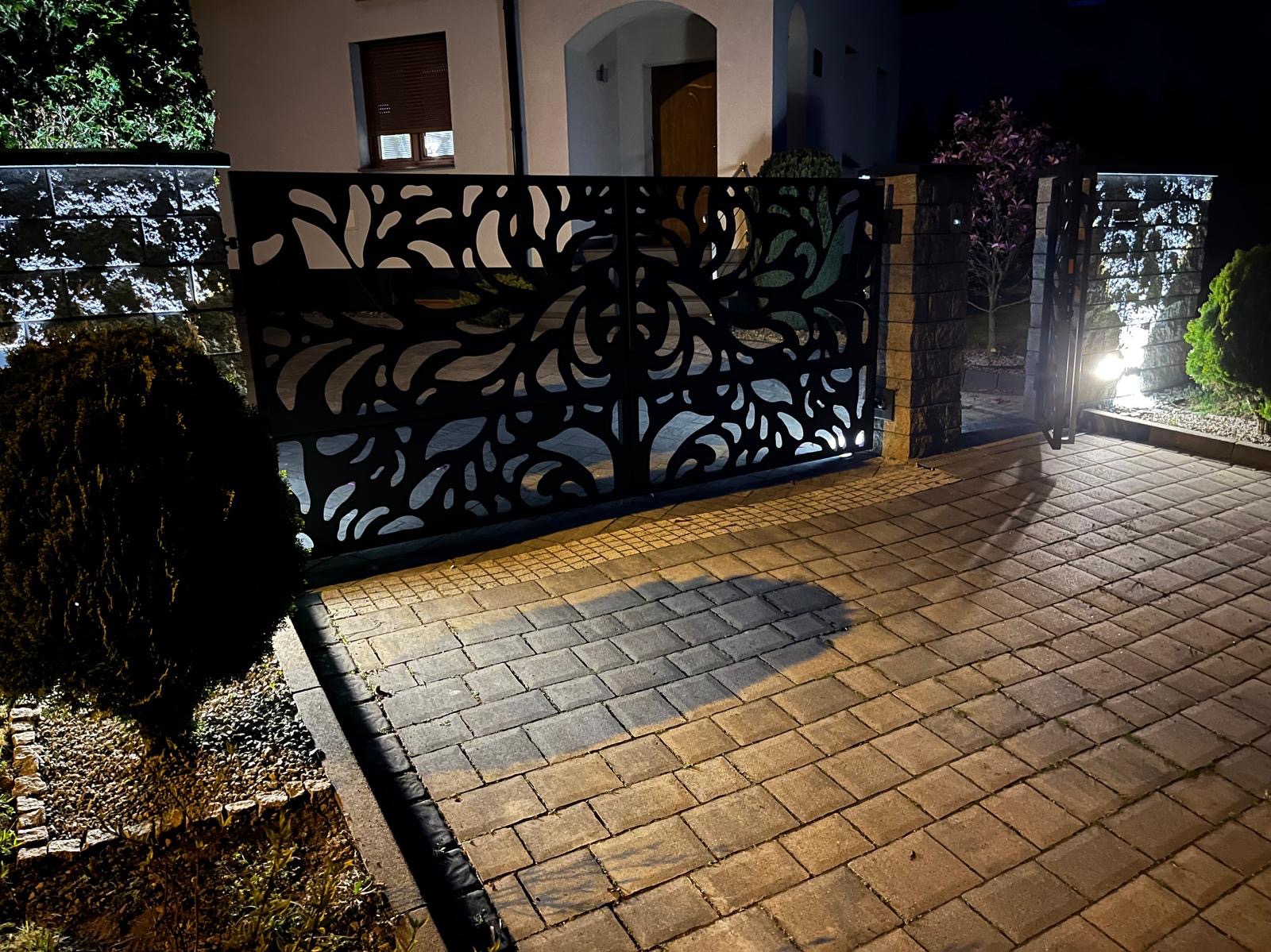 Nowoczesna brama wjazdowa z metalu, z ażurowym wzorem roślinnym, oświetlona lampami, widoczna nocą na tle domu z kostki brukowej.