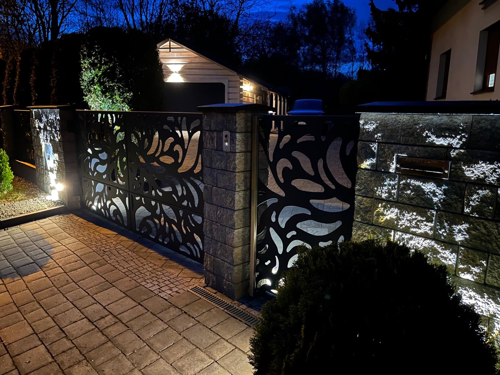 Nowoczesna brama wjazdowa z metalowymi panelami o ażurowym wzorze roślinnym, podświetlona punktowymi lampami, osadzona na murowanych słupkach z kamienia w wieczornej scenerii.