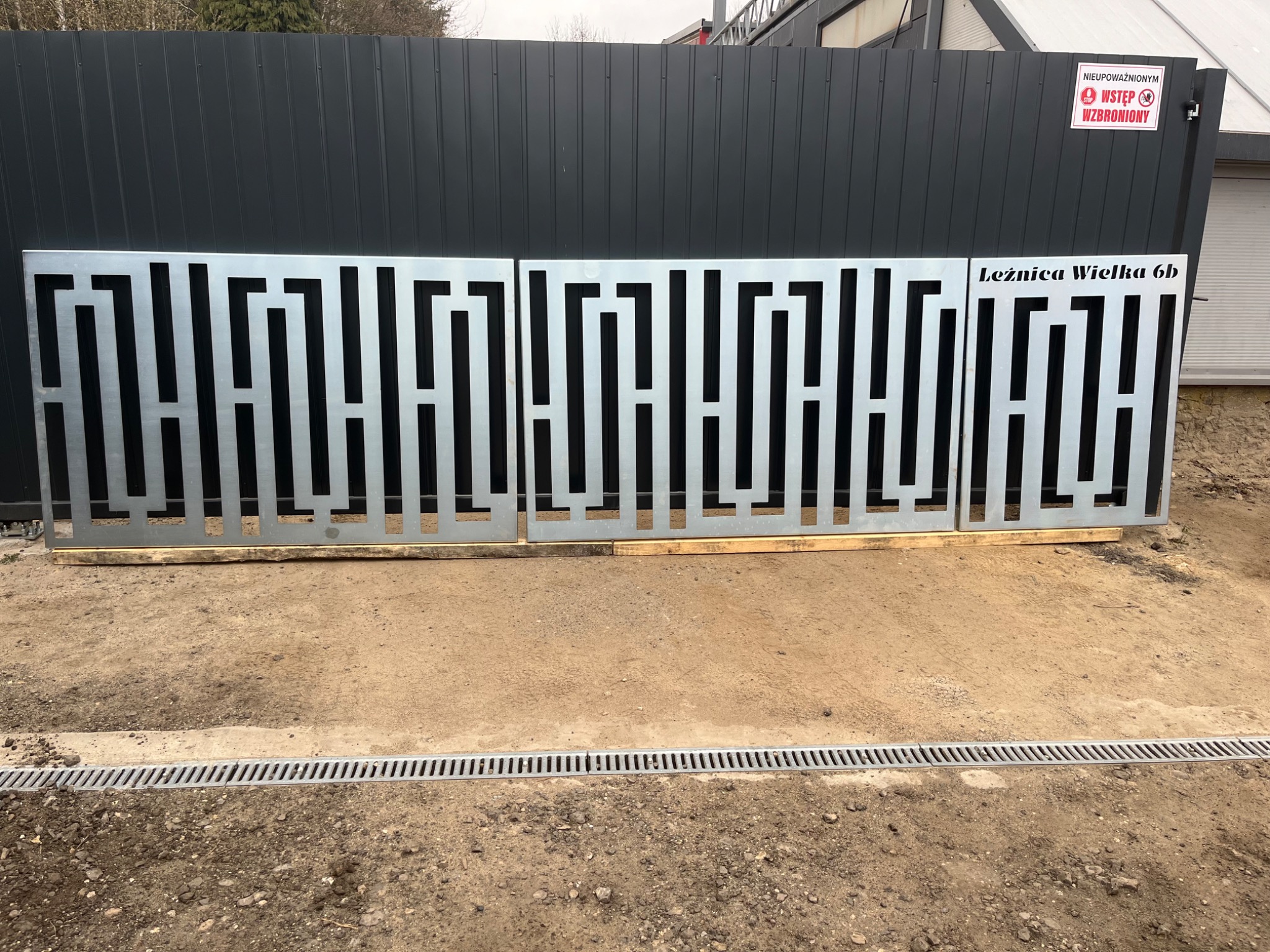 Ażurowe panele metalowe z geometrycznym wzorem labiryntu, przygotowane do montażu ogrodzenia na placu budowy, widoczny adres 'Leźnica Wielka 6b' na jednym z paneli.