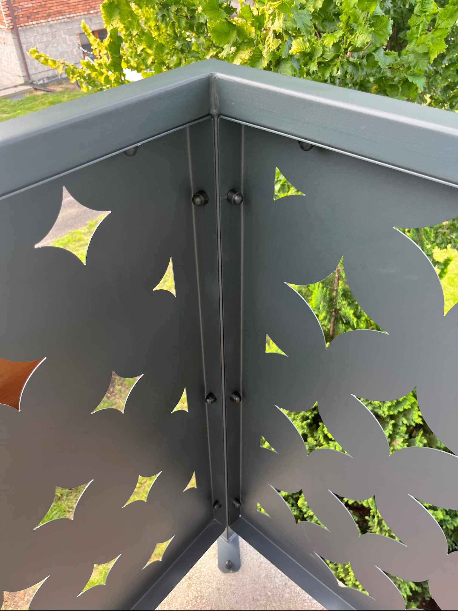 Szczegółowe ujęcie narożnika metalowego ogrodzenia z dekoracyjnymi wycięciami w kształcie liści i gwiazd, zamontowanego w Cykarzewie Północnym, z widokiem na zieleń w tle.